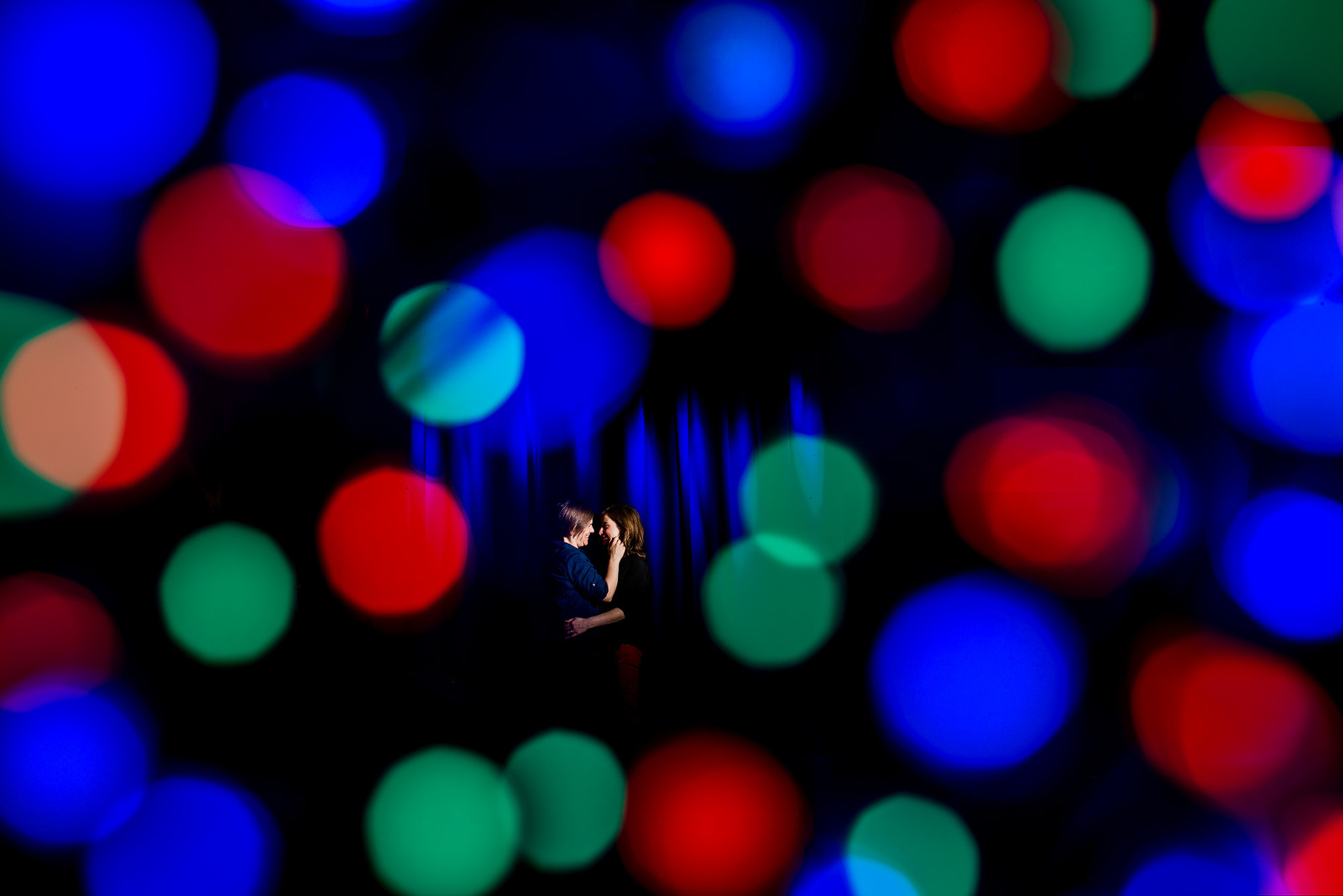 Gay couple holding each other on stage at The Masquerade with blue backdrop and colorful bokeh balls in Atlanta