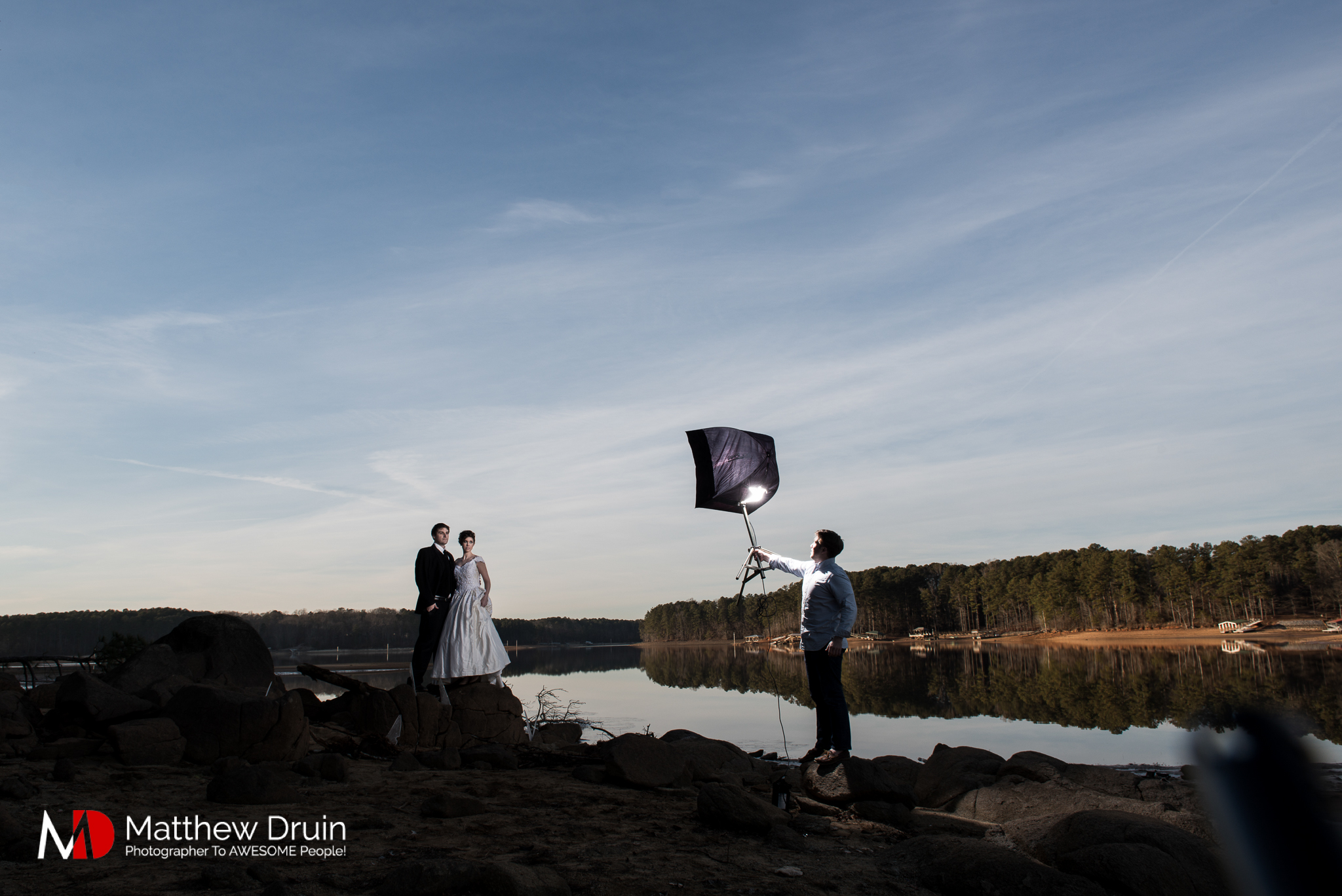 Behind the scenes of photo assistant holding a light on bride and groom Groom standing in frozen lake in Atlanta at sunset